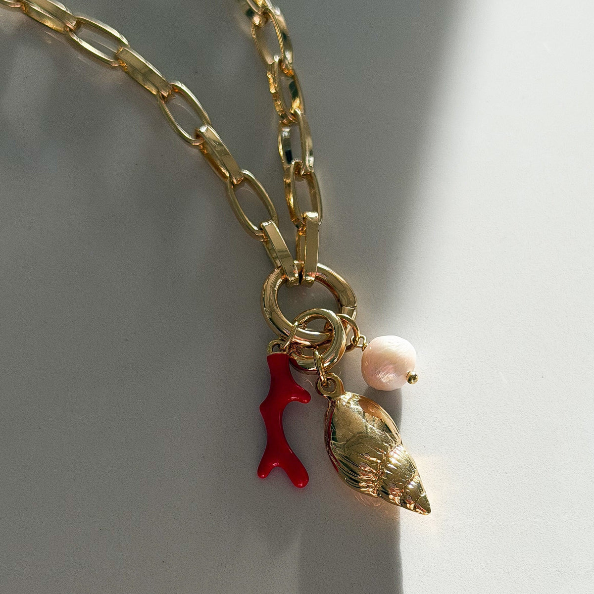 A gold chain necklace with a shell-shaped charm, a small red coral charm, and a pearl bead, photographed on a white surface with strong sunlight and shadows.
